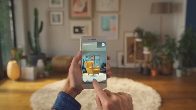 Eine Hand mit einem Smartphone, darin ein Stuhl, wie er in der echten Stube hintendran aussehen würde. Eine Hand mit einem Smartphone, darin ein Stuhl, wie er in der echten Stube hintendran aussehen würde.
