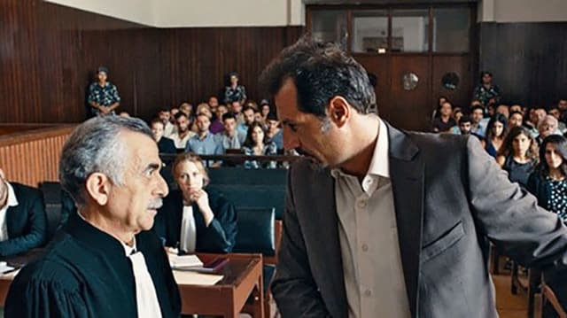 Im Gerichtssaal, Blick aus der Richtung der Richter in den Saal, Wände und Bänke aus Holz, die Reihen gut gefüllt mit Menschen. Im Vordergrund zwei Männer: Der rechts ist jünger, grosser, trägt Jacket und Hemd. Er schaut bedrohlich zum kleineren herunter, dieser trägt ein langes schwarzes Gewand mit weissem Halstuch. Im Gerichtssaal, Blick aus der Richtung der Richter in den Saal, Wände und Bänke aus Holz, die Reihen gut gefüllt mit Menschen. Im Vordergrund zwei Männer: Der rechts ist jünger, grosser, trägt Jacket und Hemd. Er schaut bedrohlich zum kleineren herunter, dieser trägt ein langes schwarzes Gewand mit weissem Halstuch.
