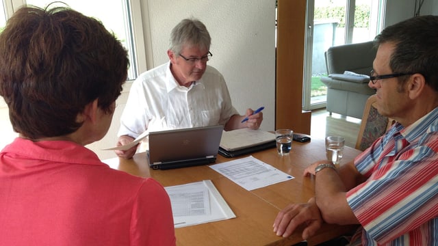 Der Versicherungsexperte sitzt vor einem Laptop und prüft ein Papier. Ein Ehepaar sitzt gegenüber und schaut gespannt. Der Versicherungsexperte sitzt vor einem Laptop und prüft ein Papier. Ein Ehepaar sitzt gegenüber und schaut gespannt.