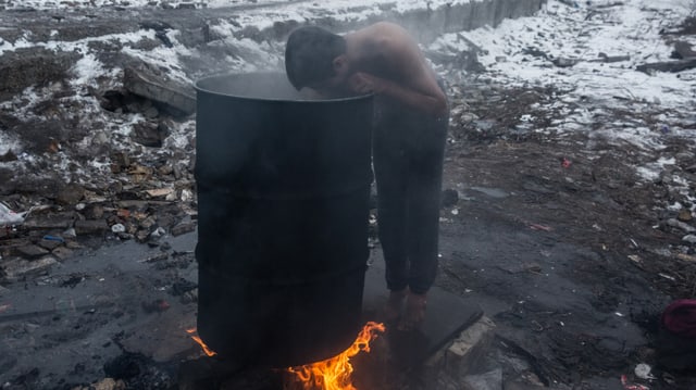 Junger Mann mit nacktem Oberkörper und barfuss wäscht sich draussen in der Kälte mit warmen Wasser aus einer Tonne, die über dem offenen Feuer erhitzt wird Junger Mann mit nacktem Oberkörper und barfuss wäscht sich draussen in der Kälte mit warmen Wasser aus einer Tonne, die über dem offenen Feuer erhitzt wird