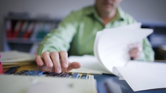 Mann blättert in Unterlagen und tippt auf einem Rechner herum Mann blättert in Unterlagen und tippt auf einem Rechner herum