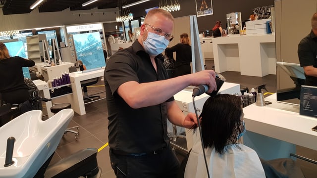 Ein Mann mit blauem Mundschutz föhnt einer Kundin die Haare. Ein Mann mit blauem Mundschutz föhnt einer Kundin die Haare.