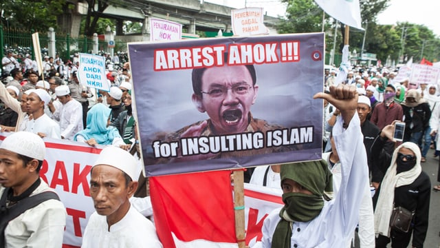 Muslime mit weissen Kappen tragen ein Transparent mit einem Foto von Basuki und der Aufschrift: «Verhaftet ihn, weil er den Islam beleidigt hat.» Muslime mit weissen Kappen tragen ein Transparent mit einem Foto von Basuki und der Aufschrift: «Verhaftet ihn, weil er den Islam beleidigt hat.»