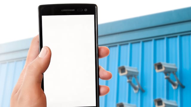 Eine Hand hält ein Handy, im Hintergrund sind vier Überwachungskameras an einer Wand montiert. Eine Hand hält ein Handy, im Hintergrund sind vier Überwachungskameras an einer Wand montiert.