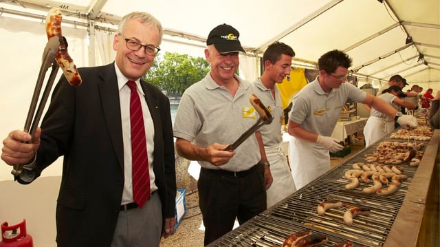 Mann mit Krawatte hält mit einer Grillzange eine Wurst in die Höhe Mann mit Krawatte hält mit einer Grillzange eine Wurst in die Höhe