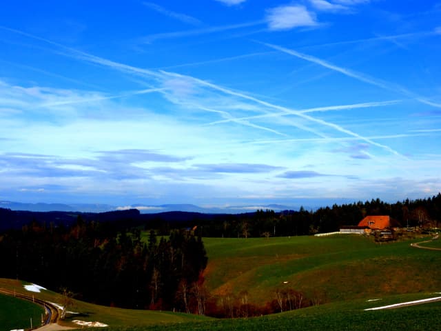 Im Vordergrund liegt eine grüne Wiese, etwas Wald und ein Bauernhaus. Der Himmel erstrahlt noch in tiefem Blau. Am Horizont erkennt man aber, dass das Wetter bald drehen wird.