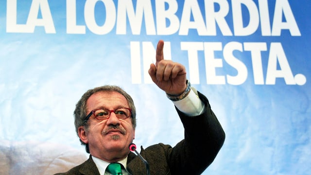 Meroni vor einem Wahlplakat, er zeigt mit dem Finger in die Luft. Meroni vor einem Wahlplakat, er zeigt mit dem Finger in die Luft.