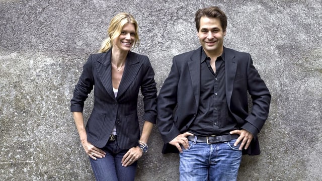Blonde Frau und braunhaariger Mann vor Fotowand blicken lachend in die Kamera. Blonde Frau und braunhaariger Mann vor Fotowand blicken lachend in die Kamera.