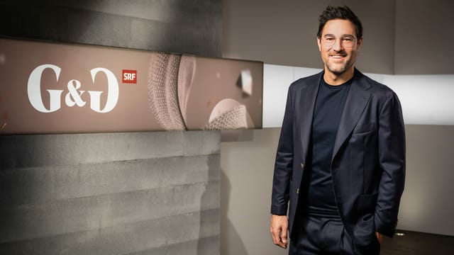 Mann mit kurzen dunklen Haaren, Brille und dunkelblauem Anzug vor einer Betonwand mit der Aufschrift «G&G» Mann mit kurzen dunklen Haaren, Brille und dunkelblauem Anzug vor einer Betonwand mit der Aufschrift «G&G»