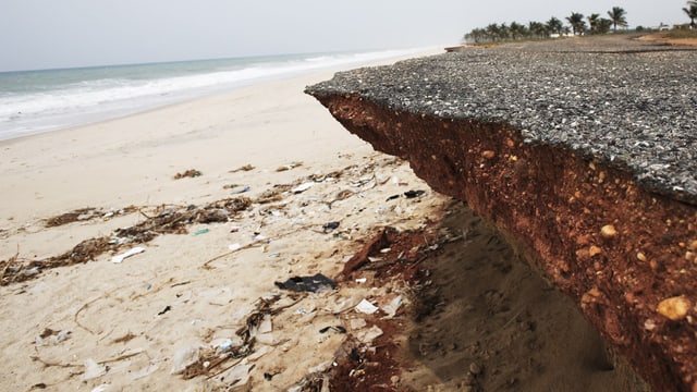 Die an den Rändern abgebrochene und unterspülte Strasse ragt über den Sandstrand. Die an den Rändern abgebrochene und unterspülte Strasse ragt über den Sandstrand.