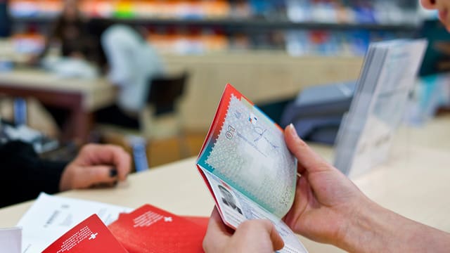 Hände einer Reisebüroangestellten halten einen geöffneten Schweizer Pass, auf dem Tisch liegen zwei weitere rote Pässe. Hände einer Reisebüroangestellten halten einen geöffneten Schweizer Pass, auf dem Tisch liegen zwei weitere rote Pässe.