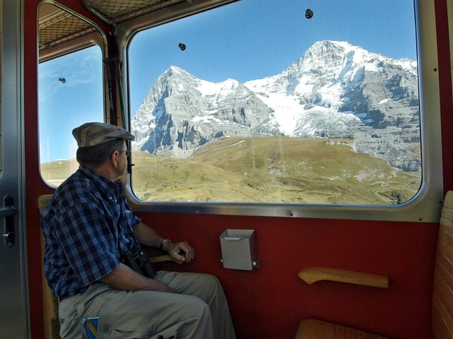 Zu sehen ist ein Mann mit blau-schwarz kariertem Hemd und Schiebermütze, wie er in der Jungfraubahn aus dem Fenster sieht. Da zu sehen ist eine grüne Bergwiese und im Hintergrund zwei verschneite Berggipfel.