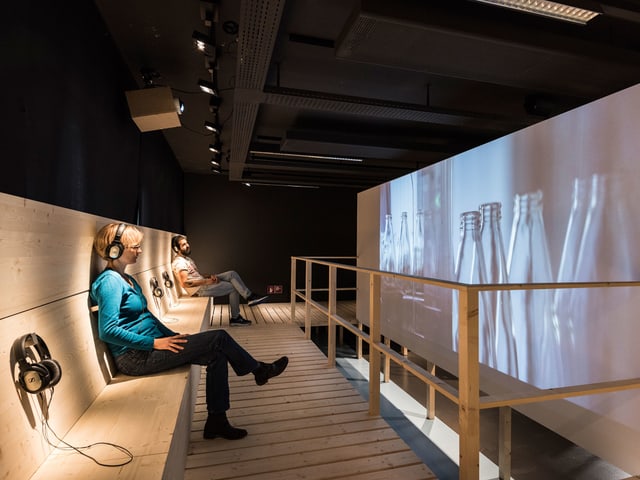 Besucher in einer Ausstellung sehen sich Flaschen auf einem Screen an. Besucher in einer Ausstellung sehen sich Flaschen auf einem Screen an.