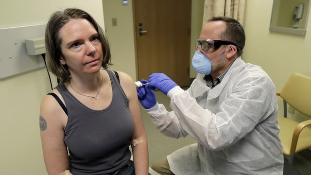 Frau sitzt auf Behandlungsliege mit Arzt daneben, der ihr einen Impfstoff in den Oberarm spritzt.