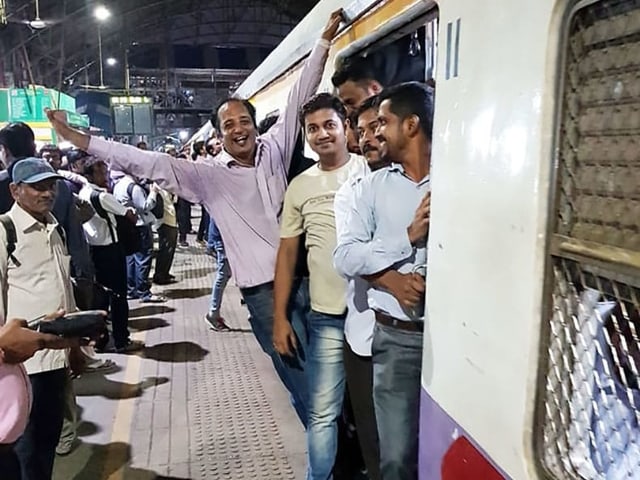 Mehrere Indische Männer stehen in der offenn Tür eines Zuges in Mumbai. Der Zug steht noch im Bahnhof. Die Männer lachen in die Kamera, einer posiert voller Freude mit einem ausgestreckten Arm, während er sich am Zug festhält. Mehrere Indische Männer stehen in der offenn Tür eines Zuges in Mumbai. Der Zug steht noch im Bahnhof. Die Männer lachen in die Kamera, einer posiert voller Freude mit einem ausgestreckten Arm, während er sich am Zug festhält.
