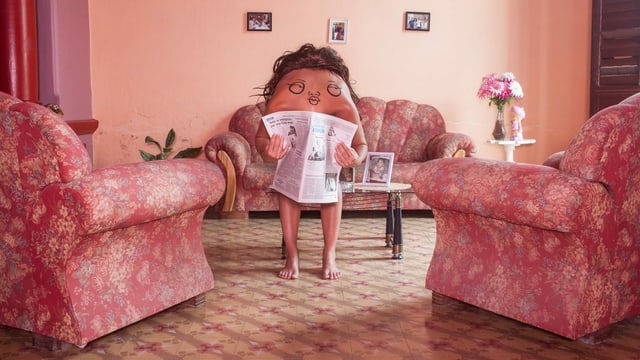 Ein Mann beugt sich im Wohnzimmer nach vorne, auf dem Rücken ein Gesicht gemalt. Er hat eine Zeitung in der Hand. Ein Mann beugt sich im Wohnzimmer nach vorne, auf dem Rücken ein Gesicht gemalt. Er hat eine Zeitung in der Hand.