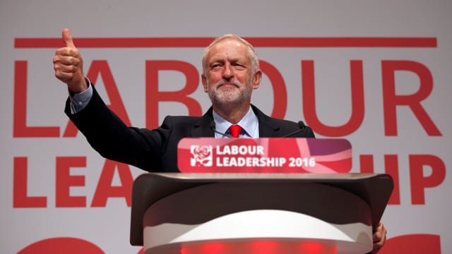 Corbyn steht hinter einem Stehpult und hält den Daumen hoch. Corbyn steht hinter einem Stehpult und hält den Daumen hoch.