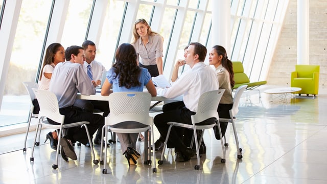 Businessleute sitzen um einen Tisch. Die Chefin steht. Businessleute sitzen um einen Tisch. Die Chefin steht.