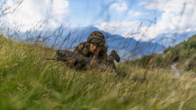 Rekrut liegt mit Waffe im Gras, dahinter Bergpanorama. Rekrut liegt mit Waffe im Gras, dahinter Bergpanorama.