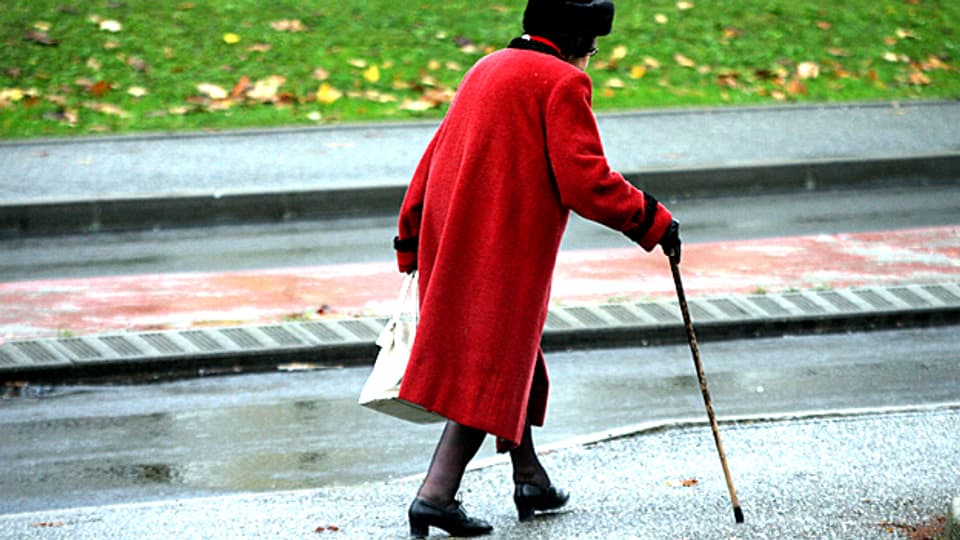 Diese Seniorin macht es genau richtig. Sie trägt einen auffällig, roten Mantel und geht auf dem Trottoir.