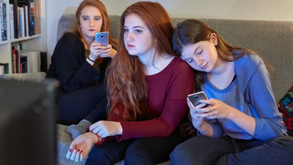Mädchen benutzen ihre Smartphones während sie im Wohnzimmer auf dem Sofa sitzend fersehen.