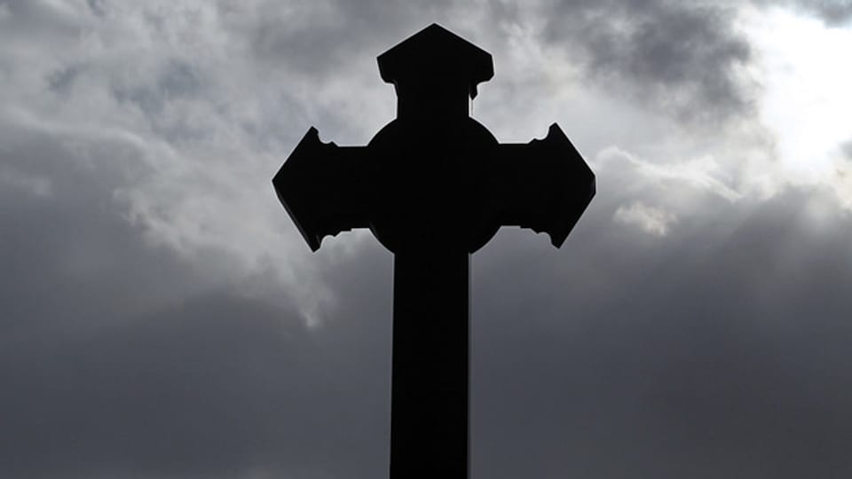 Ein Kirchenkreuz, im Hintergrund dunkle Wolken und ein Sonnenstrahl.