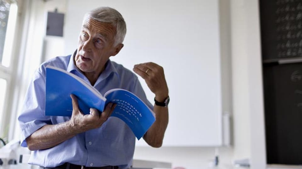 Ein älterer Lehrer hält ein Buch in der Hand und unterrichtet vor einer Klasse.