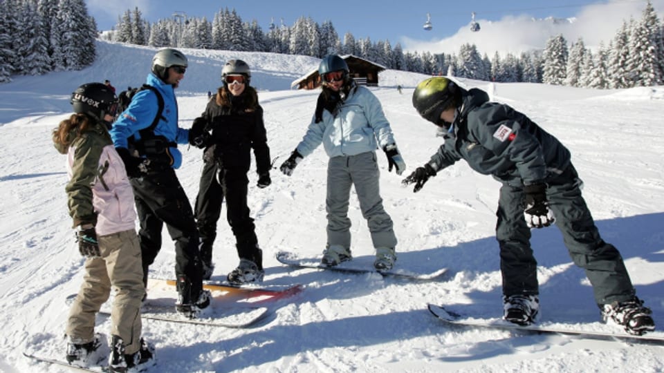 Fünf Jugendliche stehen etwas wacklig auf ihren Snowboardbrettern auf einer weissen Piste in den Bergen und lachen