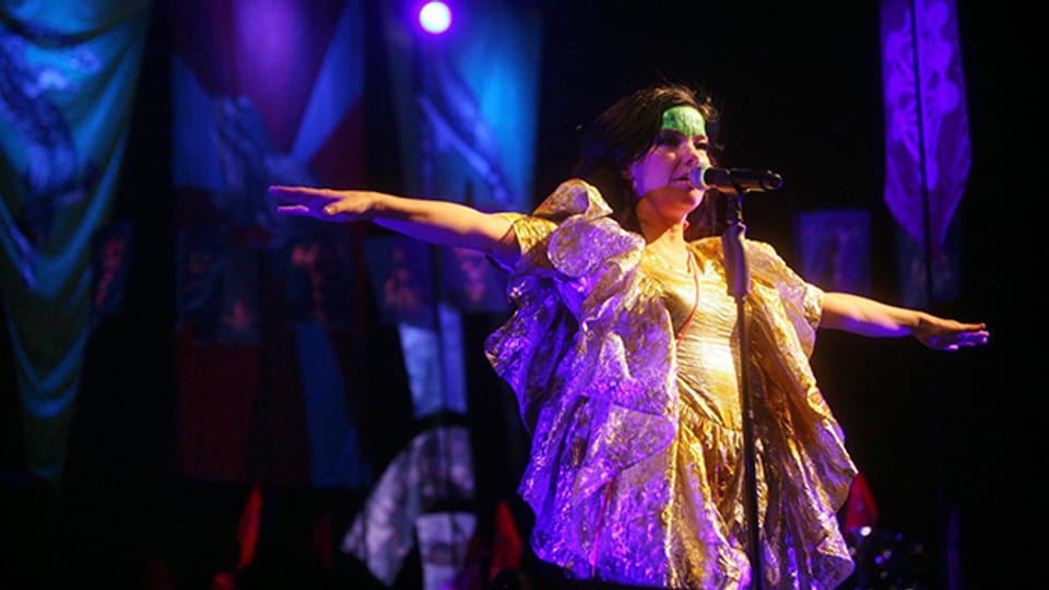 Björk mit bunt geschminktem Gesicht und ausgefallener Kleidung auf einer Bühne.