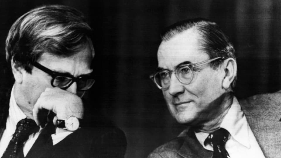 Der investigative Journalist Seymour Hersch (links) scheint dem CIA Direktor William Colby (rechts) ein Geheimnis zu zuflüstern.