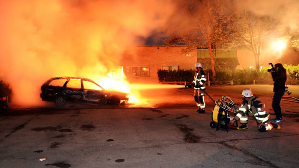 Feuerwehrmänner löschen ein brennendes Auto im Vorort Kista bei Stockholn, welches Jugendliche angezündet hatten.