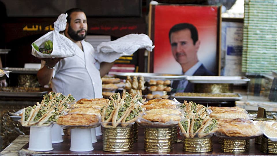 Ein Verkäufer in weissem Berufskittel verkauf syrische Süssigkeiten. Neben ihm an der Wand hängt - Bashar al-Assad