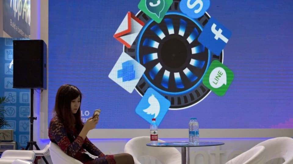 Eine Frau schaut in ihr Smartphone. Hinter ihr sind an eine Wand verschiedene Icons von Social Media Apps projiziert.