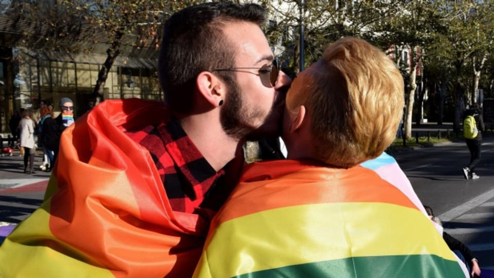 Zwei Homosexuelle küssen sich eingewickelt in eine Regenbogenfahne.