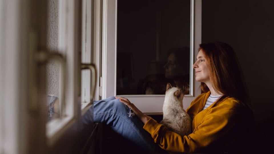 Symbolbild. Junge Frau sitzt mit ihrer Katze vor dem Fenster in ihrer Wohnung.