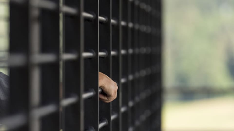Symbolbild. Eine Hand ragt zwischen Gefängnis-Gitterstäben heraus.