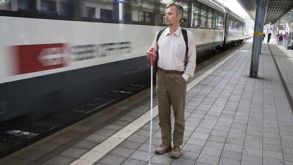 Ein blinder Mann steht auf einem Zugs-Perron.