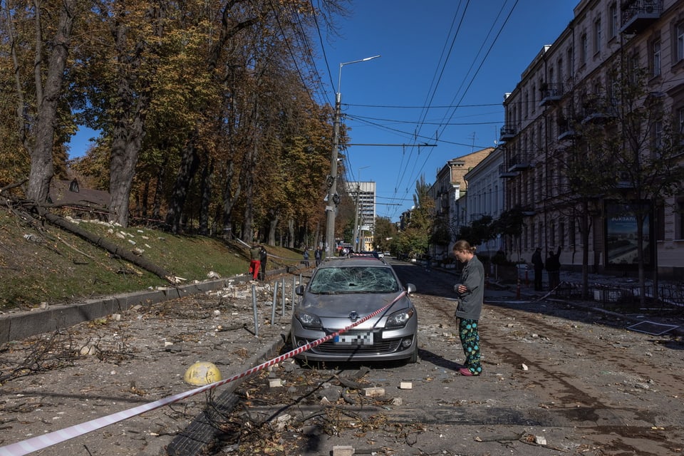 Ein kaputtes Auto auf einer Strasse in Kiew. Der Boden ist übersäht von Schutt, daneben steht ein junger Mann. Die Sonne scheint