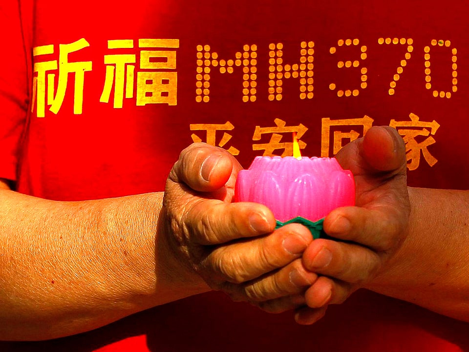 Nahaufnahme eines Mannes in einem roten MH370-Trauer-T-Shirt hält eine brennende Trauerkerze vor seinem Körper.