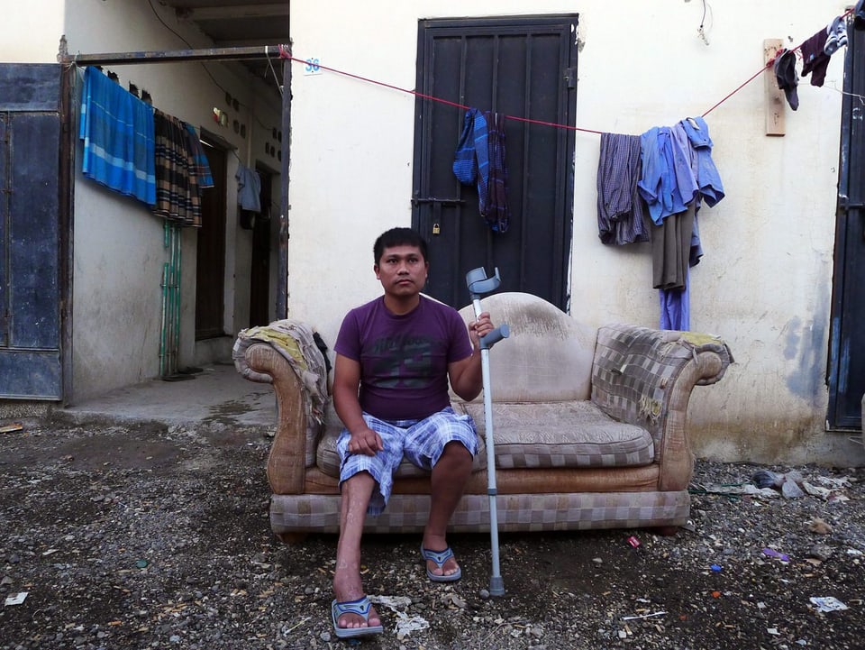 Ein Mann sitzt vor einer ärmlichen Behausung auf einem Sofa, in der einen Hand eine Krücke.