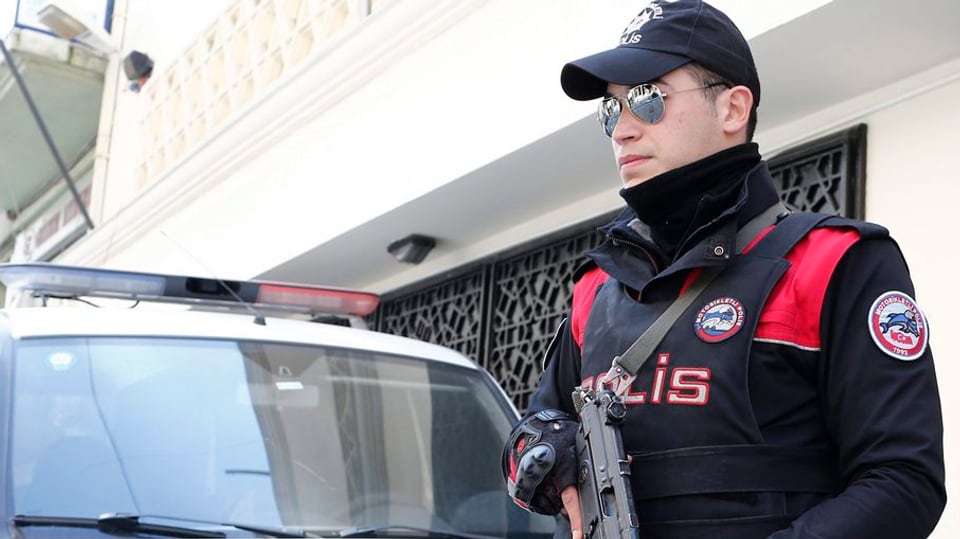Türkischer Polizist mit Maschinenpistole steht Wache vor einem Haus, hinter ihm ein Polizeifahrzeug