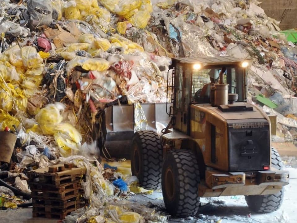 Ein Bagger hebt vor einem grossen Abfall-Haufen Kunststoffabfälle in die Höhe.