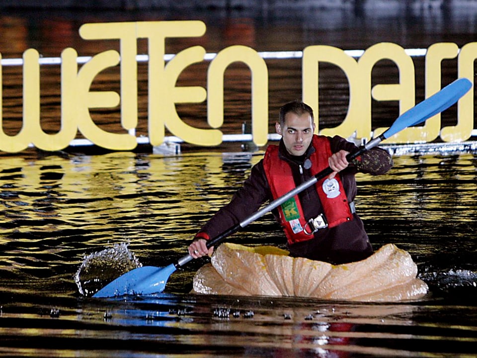 Mann paddelt in einem grossen Kürbis über einen See.