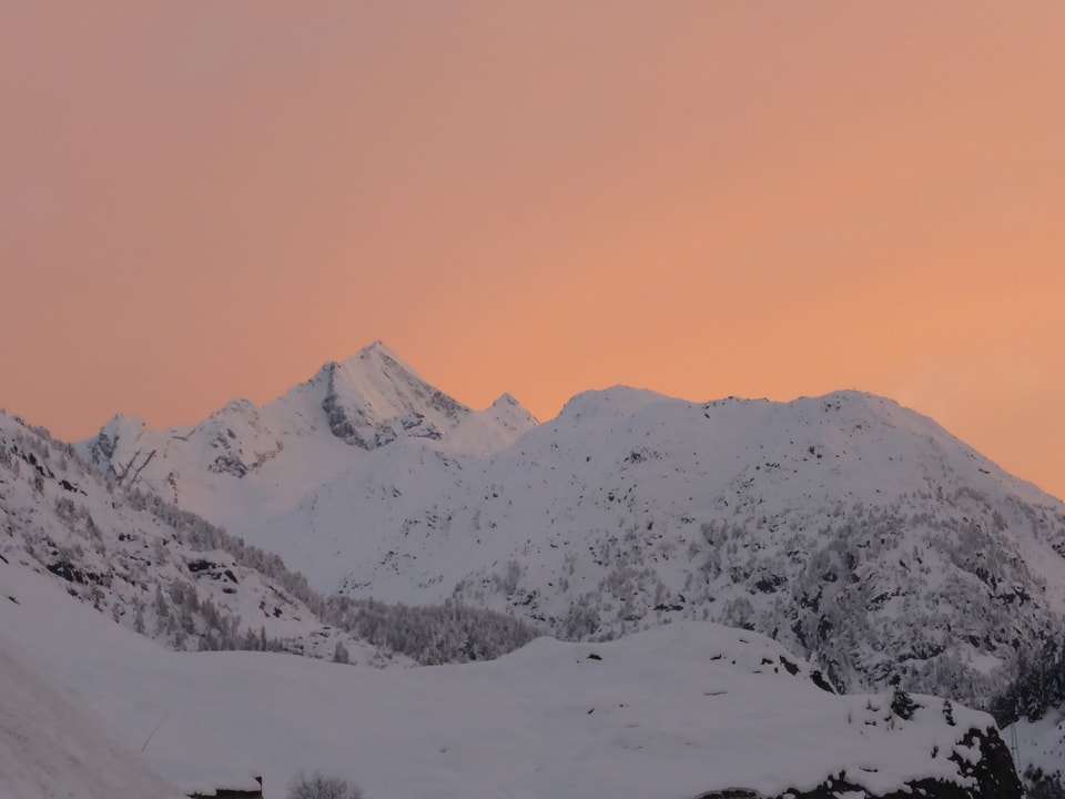 Im Vordergrund steht eine tiefverschneite Bergkette. Der Himmel über der Bergkette ist zwar noch bewölkt. Die Wolkendecke leuchtet jedoch in kitschigen Farben, die von Orange bis Rosarot reichen.