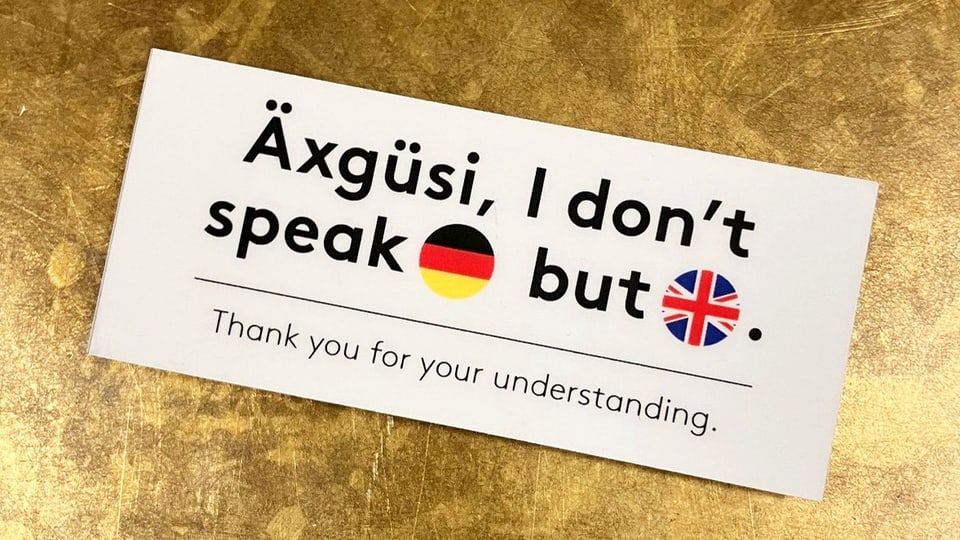 Auf einem kleinen Schild steht «Äxgüsi, I don't speak German, but English. Thank you for your understanding.»