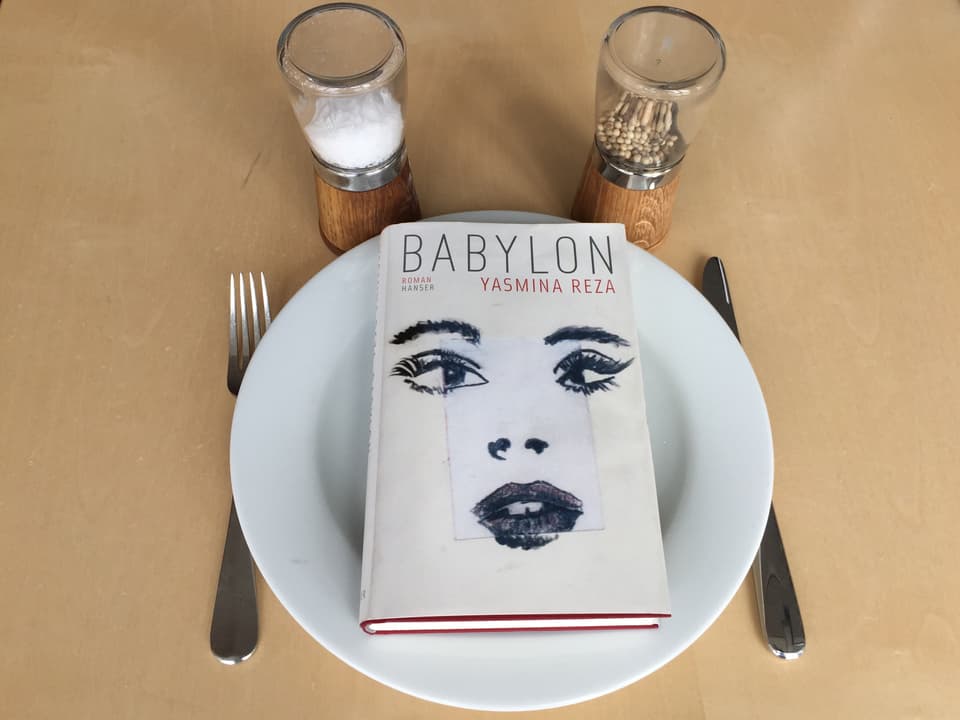 Der Roman «Babylon» von Yasmina Reza liegt auf einem weissen Teller, Messer und Gabel daneben. Oberhalb steht eine Pfeffer- und Salzmühle