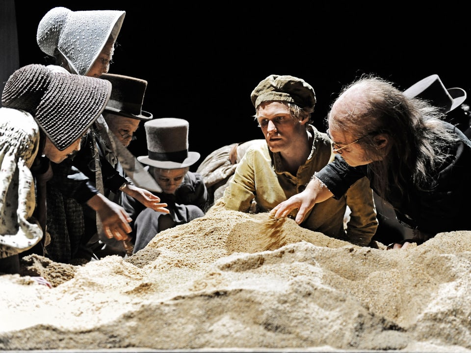 Mehrere kleinwüchsige, altmodisch gekleidete Schauspieler umringen den Sandhaufen mir Jirka Zett.