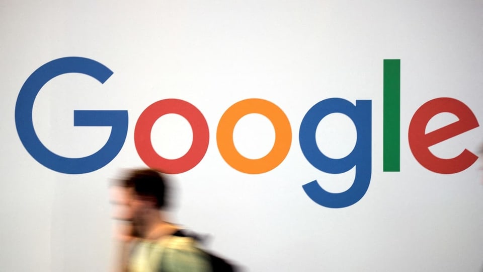 Ein telefonierender Mann läuft nach links weg vor einem Google-Logo auf einer weissen Wand.