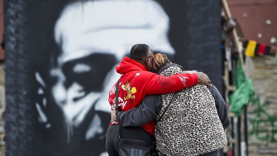 Zwei Menschen umarmen sich vor einem Wandgemälde, das George Floyd zeigt.