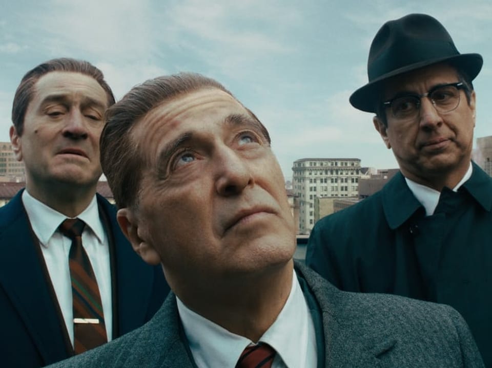 Auf dem Bild sind von links nach rechts Robert de Niro, Al Pacino und Ray Romano zu sehen. Das Bild stammt aus dem Film "The Irishman". Die Männer tragen alle Anzüge im Stil der 40er-Jahre. Ray trägt ausserdem einen schwarzen Hut.
